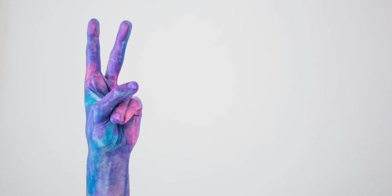 Vividly painted hand making a peace sign against a plain background.